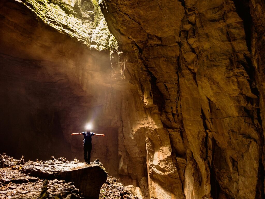 A man stands with arms spread inside a vast cave, illuminated by natural light beams in Cuetzalan, Mexico.
